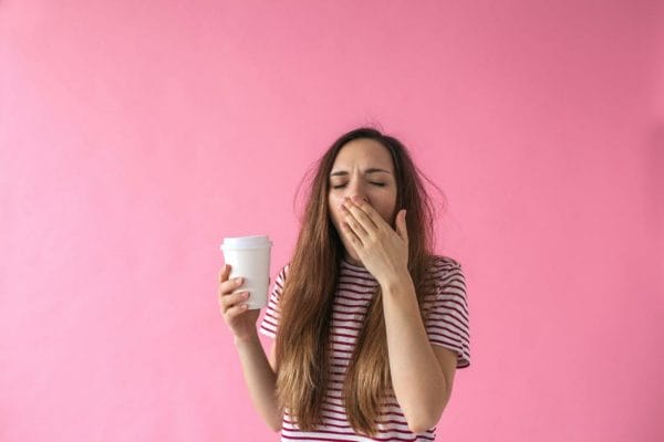 Female student yawning with coffee in hand on pink background